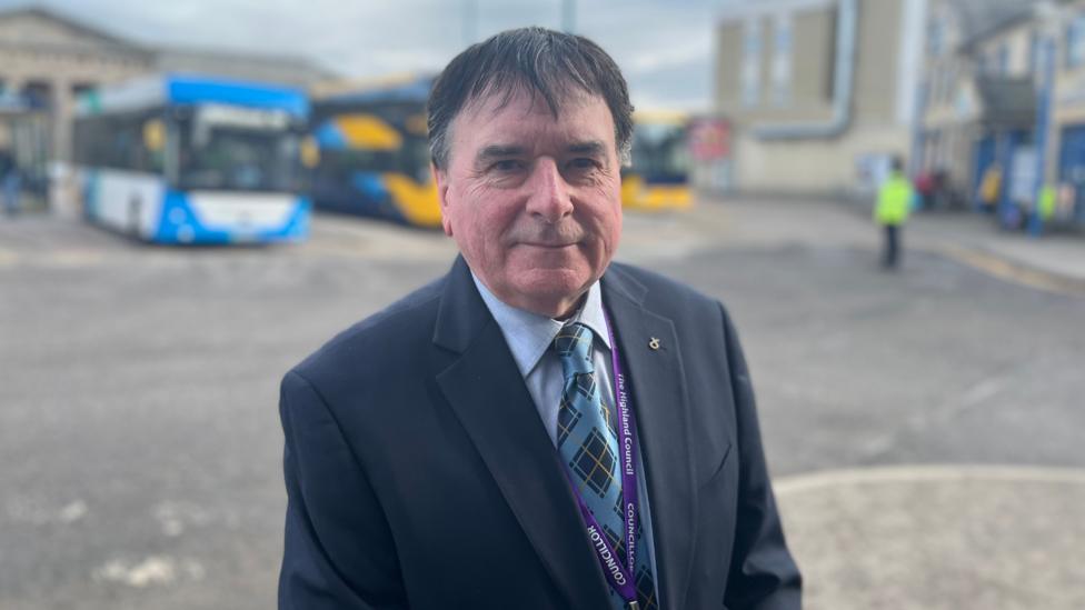 A man in a navy suit and blue tie stands in front of a blurred background where buses can be seen.