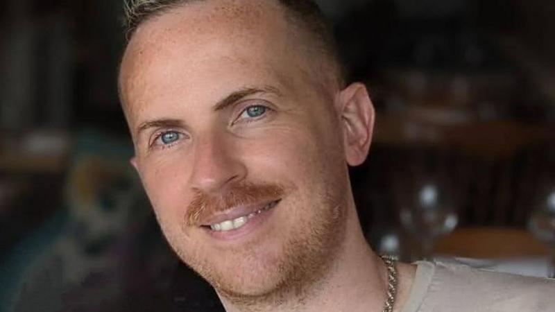Steven Elliott is smiling at the camera. He has a moustache and a light beard and is wearing a thick chain around his neck