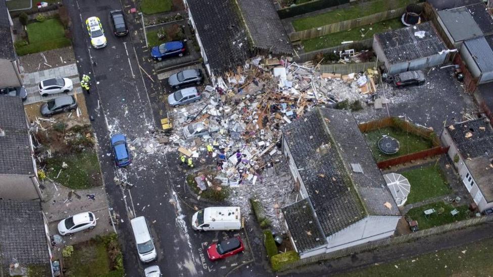 Aerial view of a partially collapsed house surrounded by rubble, with emergency workers and vehicles at the scene on a residential street.