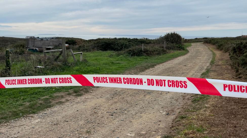 A wide, open coastal path or headland is shown under a heavy grey, overcast sky, with scrubby bushes and rough grass on either side of a dirt track. Red and white tape marked "POLICE INNER CORDON - DO NOT CROSS" has been stretched across the path.