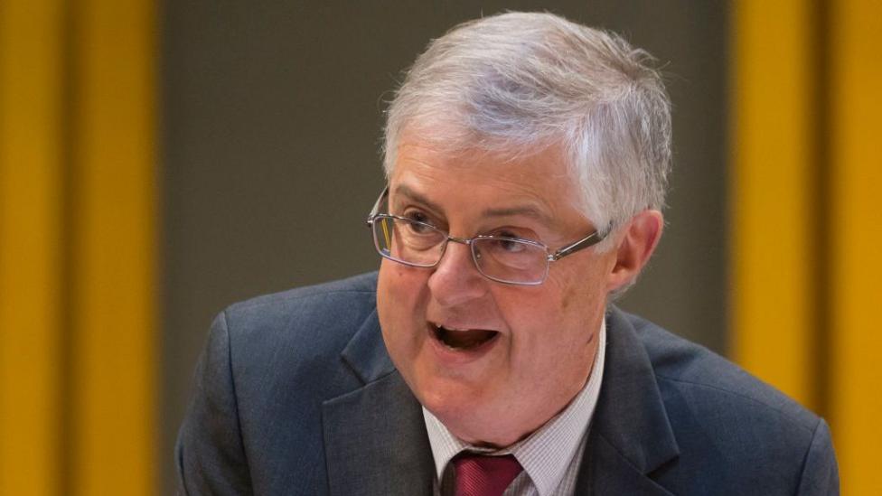 Mark Drakeford is an older man with light grey hair, glasses, wearing a dark grey suit and red tie.
