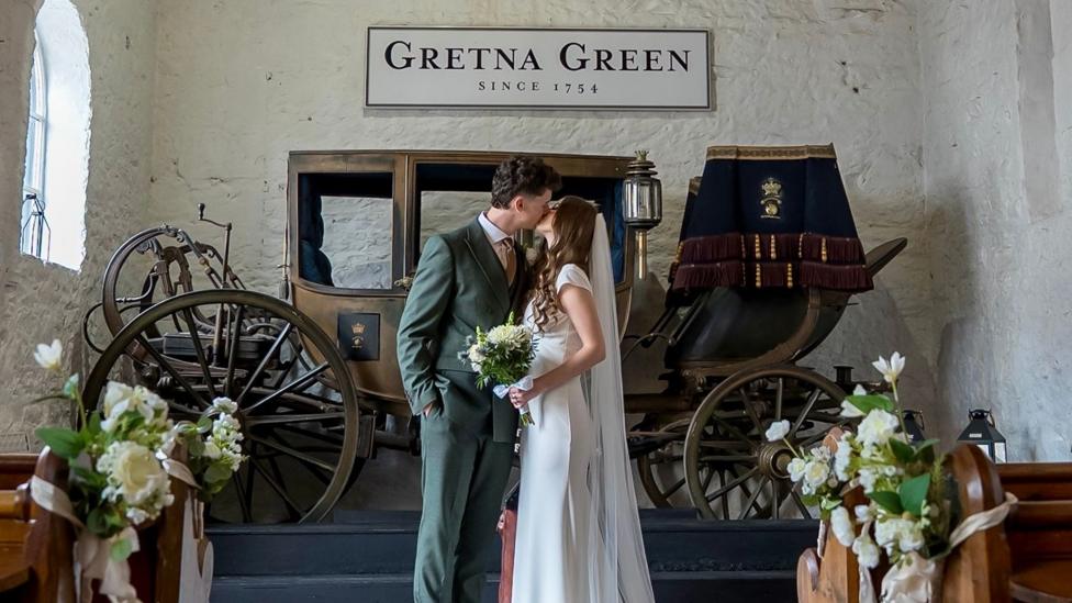 Patryk and Angelika, a young couple, kiss at the alter. Above them reads a sign "Gretna Green since 1754." The man wears a green suit and the bride wears white, with a white veil and is holding a bouquet of white flowers.