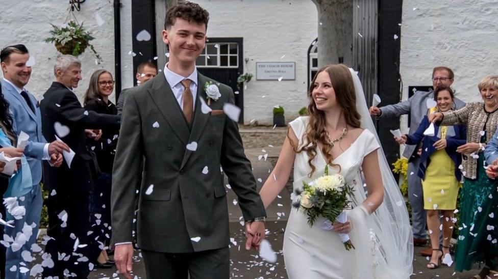 The couple walk through confetti thrown by their family members as they walk out of the hall. They both smile. Angelika looks at Patryk as he looks down the camera.