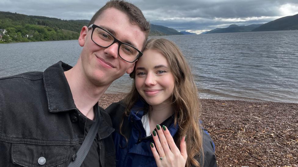 The couple stand on the banks of Loch Ness, while taking a selfie. Behind them is the glen in which the Loch sits. The smile to the camera while Angelika shows off her engagement ring.
