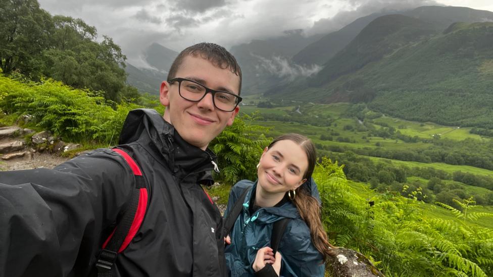 The couple are on a hike, behind them is a backdrop of green hills, fields and ferns. The pair look wet and wear rain coats and back packs. They are both smiling.