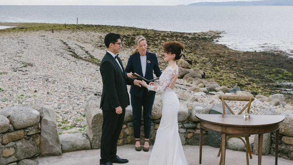 Chihon and Ana Maria stand with an officiant as they say their vows. Behind them is a stony beach with sea and hills in the distance. Chihon has black hair and wears a black suit, across from him, Ana Maria has brown hair in an updo and wears a white lace wedding dress.