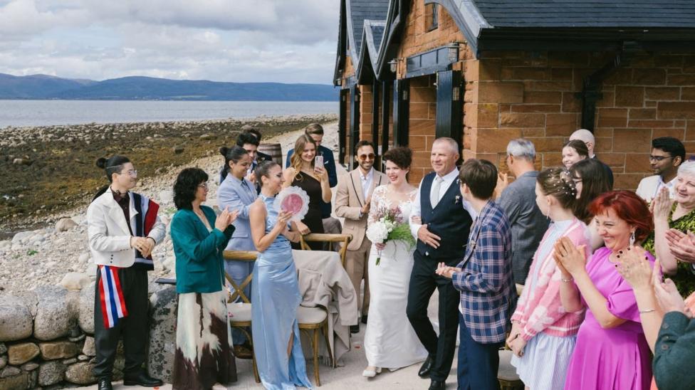 Ana Maria walks down the aisle between a sea of friends and family. She holds a bouquet in one hand and has joined arms with an older gentleman, presumably her father. Behind her guests is the coastline and distant hills.