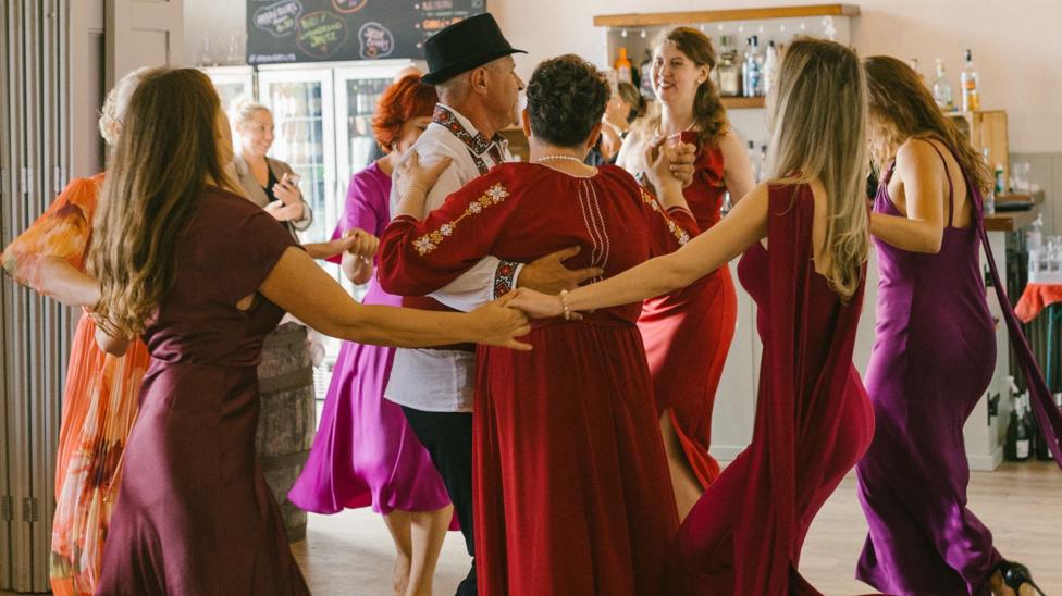 An older couple dressed in traditional Moldovan attire stand in the centre of a ceilidh circle. The woman wears a long red dress that has long sleeves with embroidery down the arms. The man wears a black short brimmed hat and a white shirt that is embroidered down the collar and round the cuffs of the sleeves. The woman dancing in a circle around them wear dresses of red and purples.