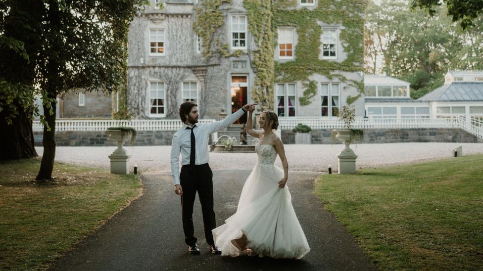 A bride and her husband stand in a castle driveway enclosed by trees. Behind them is a doorway to a castle. He is holding her hand in the air as if to turn her in a dance. It looks to be the early hours of the morning.