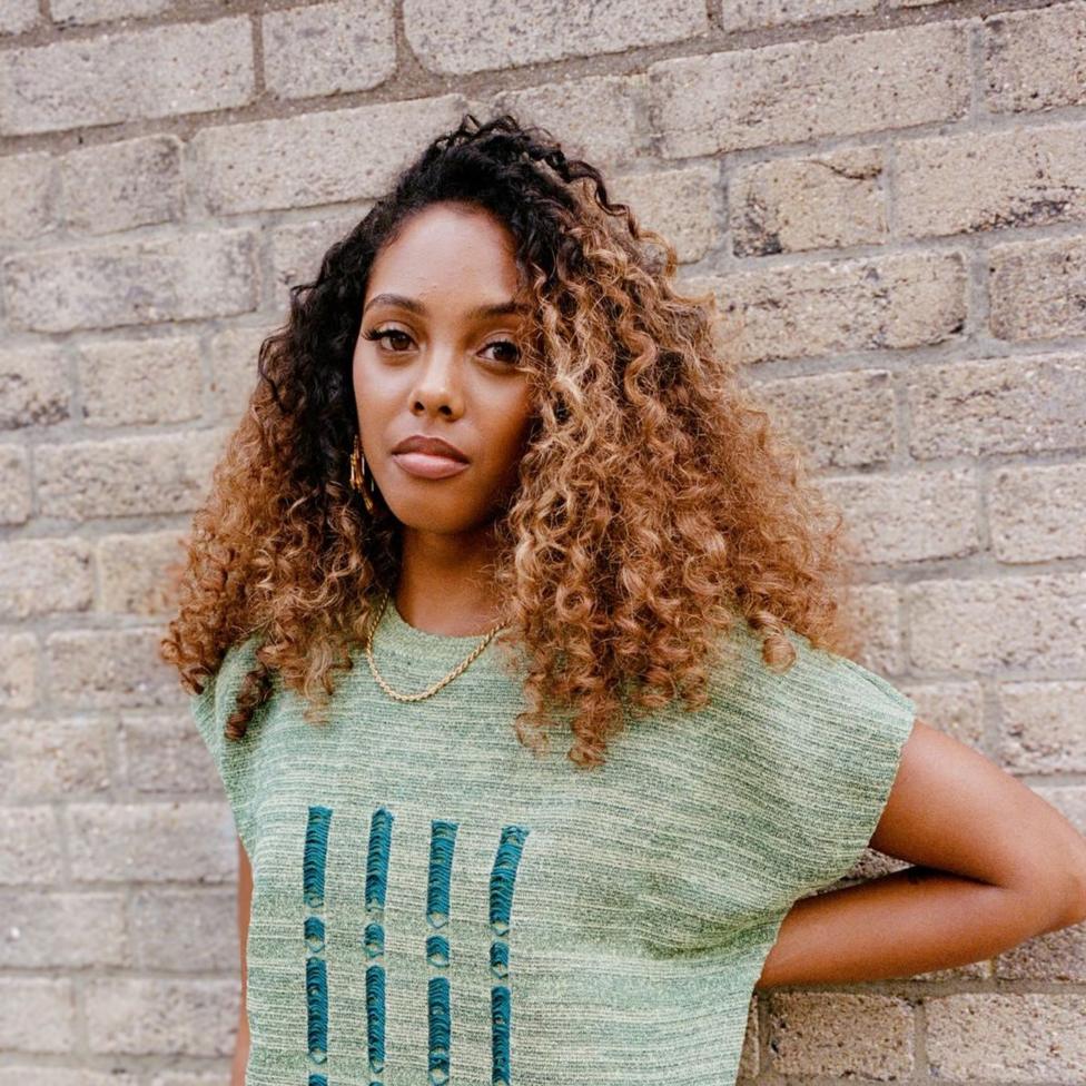 Lola Baird pictured standing by a brick wall. She has highlighted long dark afro hair and she is wearing a green, knitted, sleeveless top