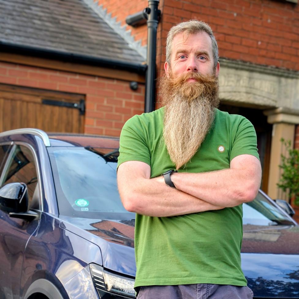 Mark Quigg, a man with a long beard in a green T-shirt and grey shorts, stands in front of a dark blue Renault, which is parked on a driveway in front of a house
