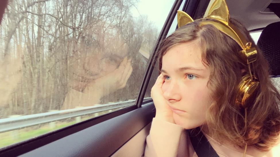A girl sits in the backseat of a car. She is resting her head in her hands and is wearing gold headphones with animal ears. She looks unhappy or bored.