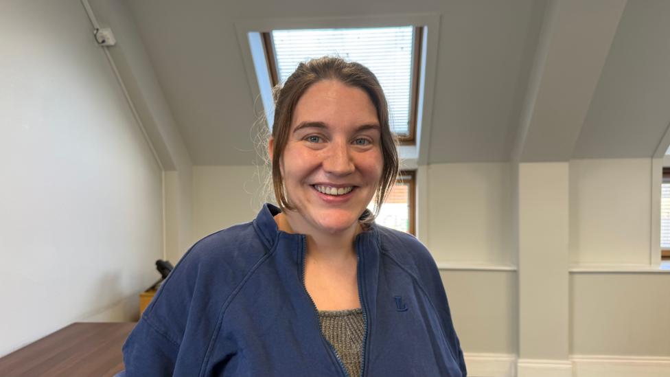 A woman with brown hair smiling at the camera. She is wearing a blue fleece. The walls behind her are white, and there is a sky-light.