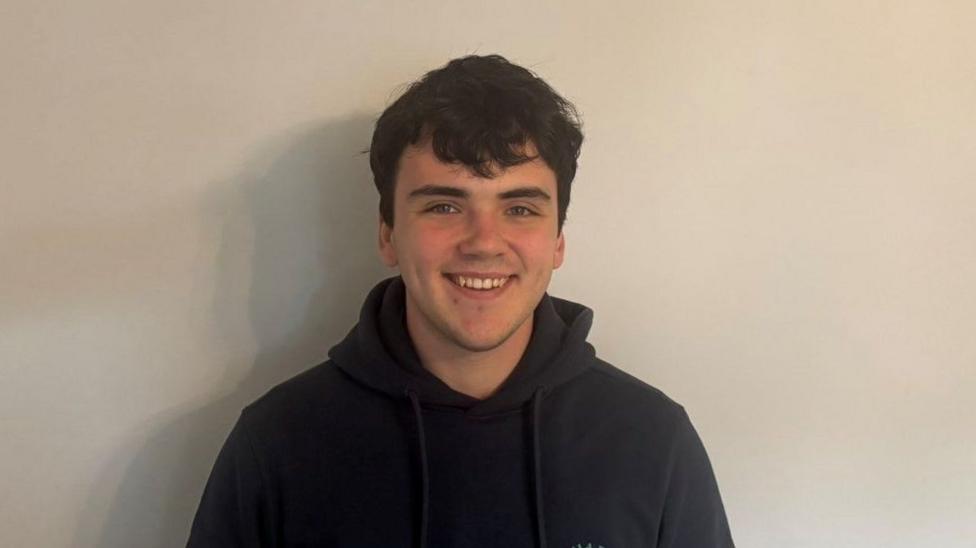 A man with black hair smiles at the camera. He is wearing a black hoodie and standing in front of a plain white wall.