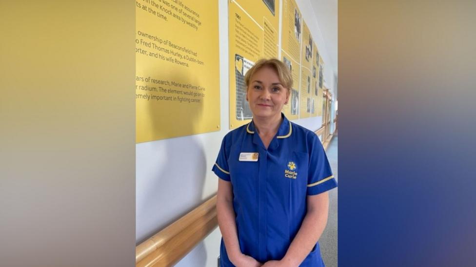 A woman with blonde hair smiles at the camera. She is wearing dark-blue hospital scrubs, which have the words 'Marie Curie' written on them in yellow. She is standing next to a wall with yellow posters.