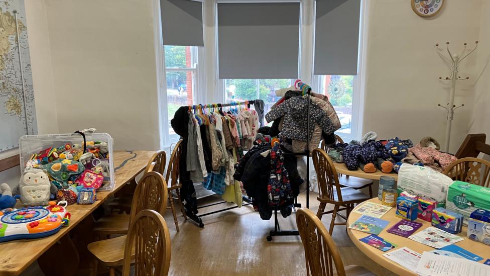 A room filled with various baby and children's items. There are small clothes rails at the back with children's clothes on, and there are wooden tables with toys, clothes, and items for parents - such as sanitary products, nappies and baby formula - on.