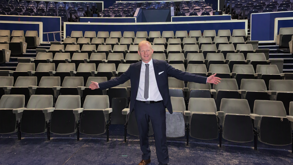A man in a suit stands with his arms spread out wide in front of row upon row of cinema seats