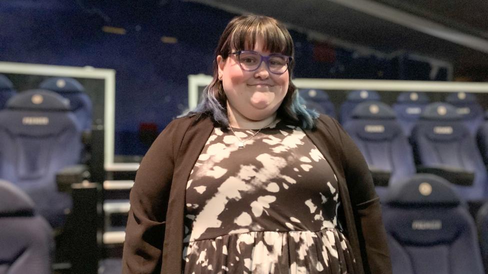 A young woman with brown hair with blue streaks in a black and white dress with a black cardigan over it. She is wearing blue glasses and standing in front of some cinema chairs.