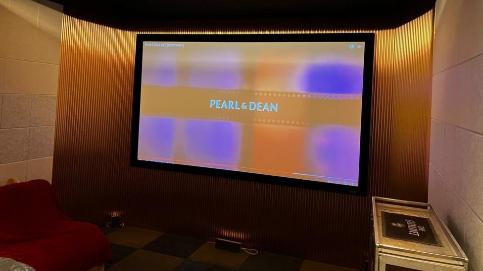 A home cinema set up with a large screen showing Pearl & Dean names and comfy seats around it