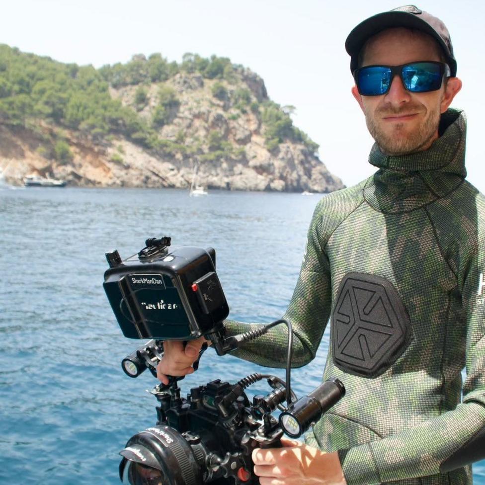 A man in a wetsuit, baseball cap and sunglasses. he is holding a camera. Behind him you can see the sea, small boats and rocky coastline.