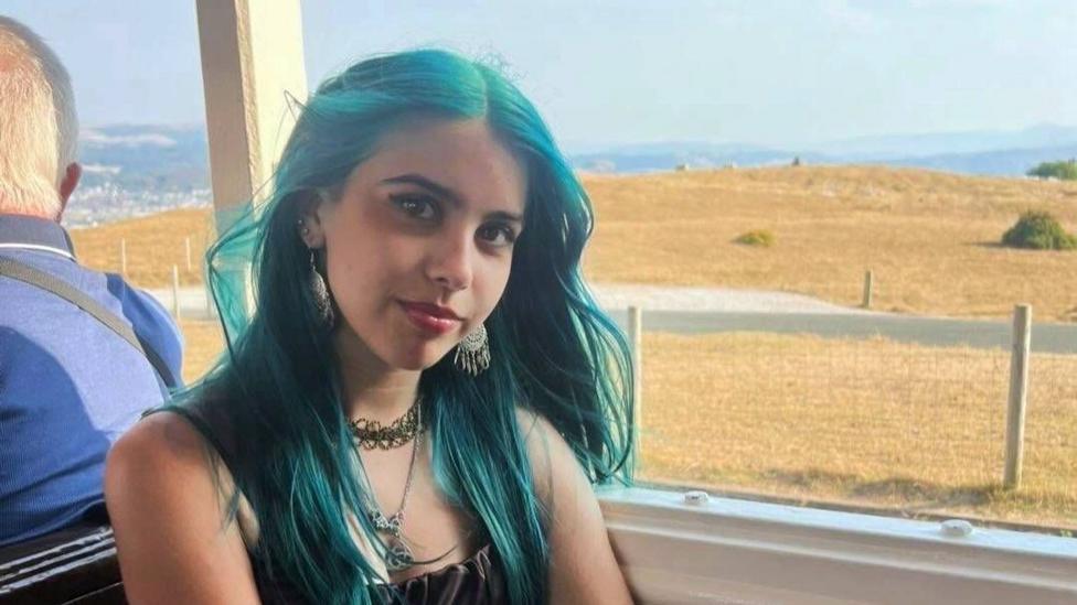 Mia looks at the camera, she has long blue hair and is wearing a black top. She seems to be on a bus, the view out of the window is a field with brown grass and mountains in the background.