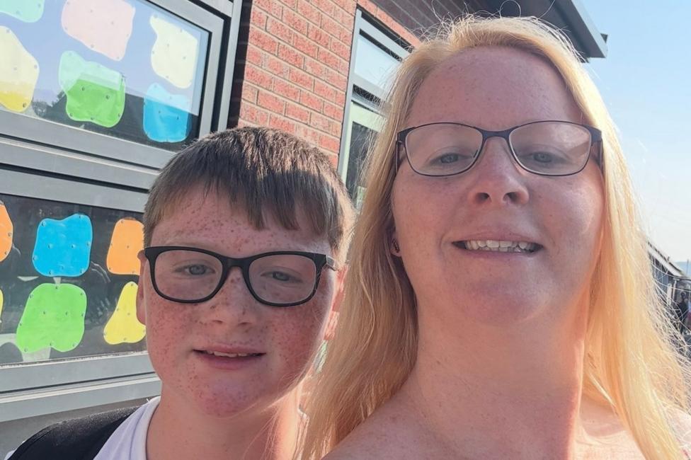 A selfie taken outside a school building on a bright sunny day. Two people stand close together in the foreground, looking towards the camera. Behind them is a brick wall and a row of windows decorated with colourful paper shapes, while clear blue sky is visible above.