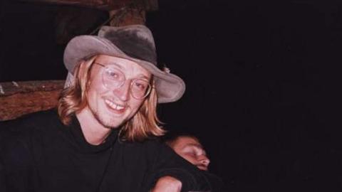 A young man with mid length blond hair, smiling at the camera. He's wearing a big cowboy style hat, a black long-sleeved top and clear glasses.