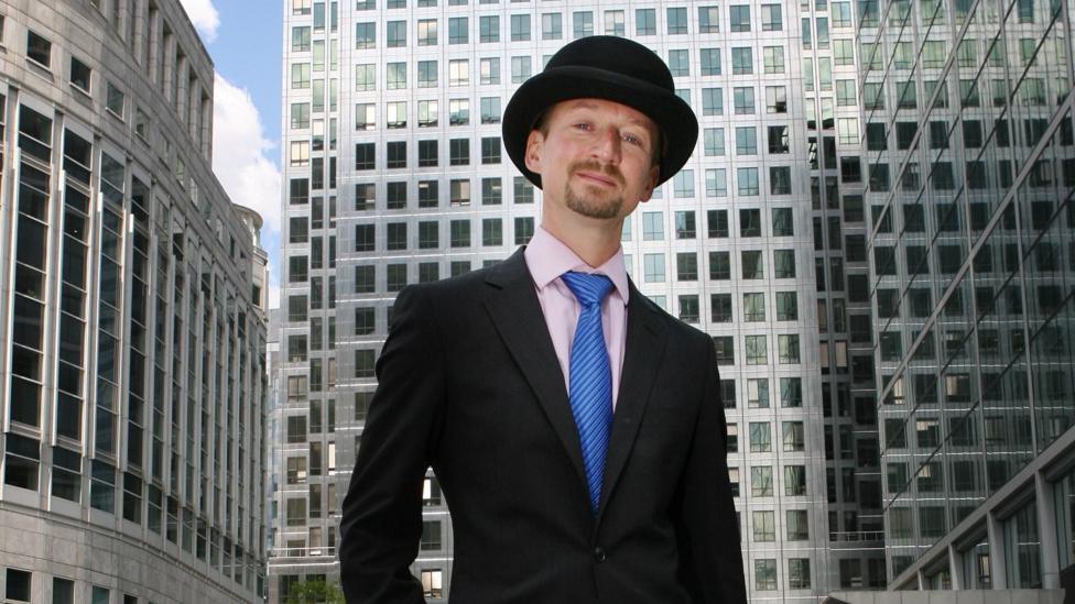 A man in a black suit and top hat. He has a pink shirt and stripy blue tie. He also has a short beard and is surrounded by office buildings.