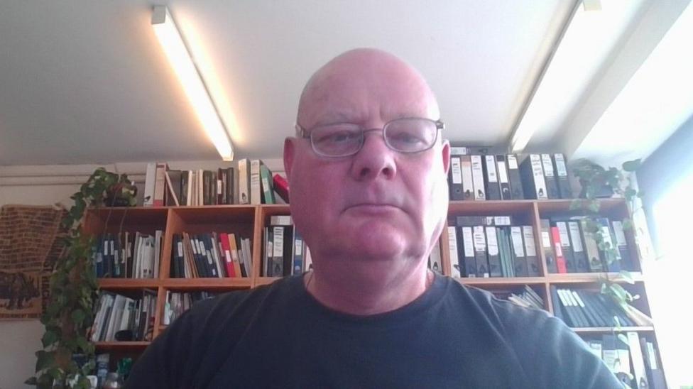A bald man looking directly at the camera, wearing a black t-shirt and glasses. He has a shelf full of books behind him.