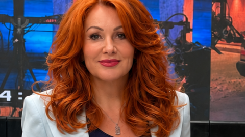 A woman with big ginger hair looking at the camera. She has a full face of makeup on and wearing a pale blue suit jacket.