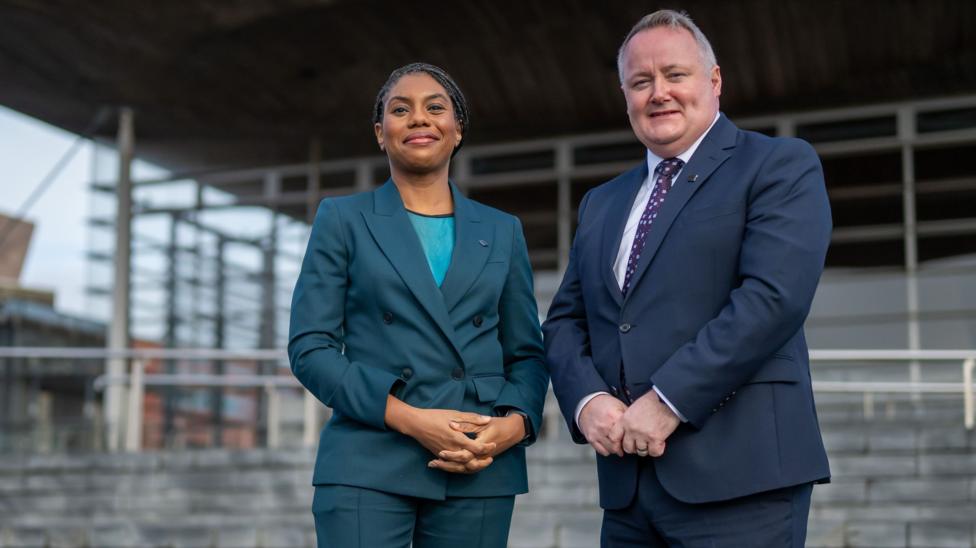 Kemi Badenoch wearing a teal suit stood next to Darren Millar. He is wearing a navy suit with a purple patterned tie.