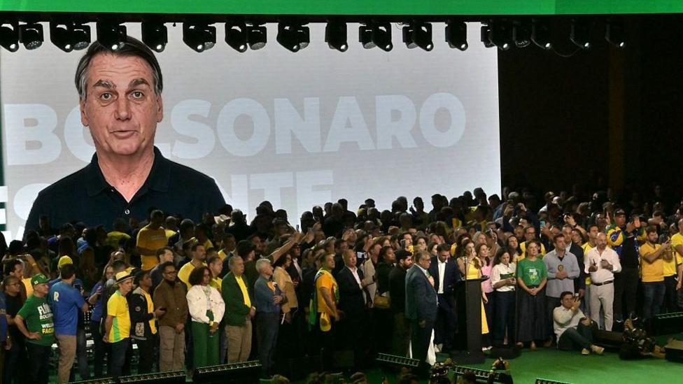 An AI-generated video of former president Jair Bolsonaro is screened during the launch of the presidential candidacy of his son, Flávio Bolsonaro, at the Liberal Party convention, in São Paulo on 25 July, 2026. The image of Jair Bolsonaro, wearing a black polo shirt, appears against a grey and white background with the words "Bolsonaro presidente" emblazoned across it. In the foreground, Flávio Bolsonaro can be seen standing in front of a lectern. He is surrounded by dozens of his supporters, some of whom are wearing Brazilian football shirts.