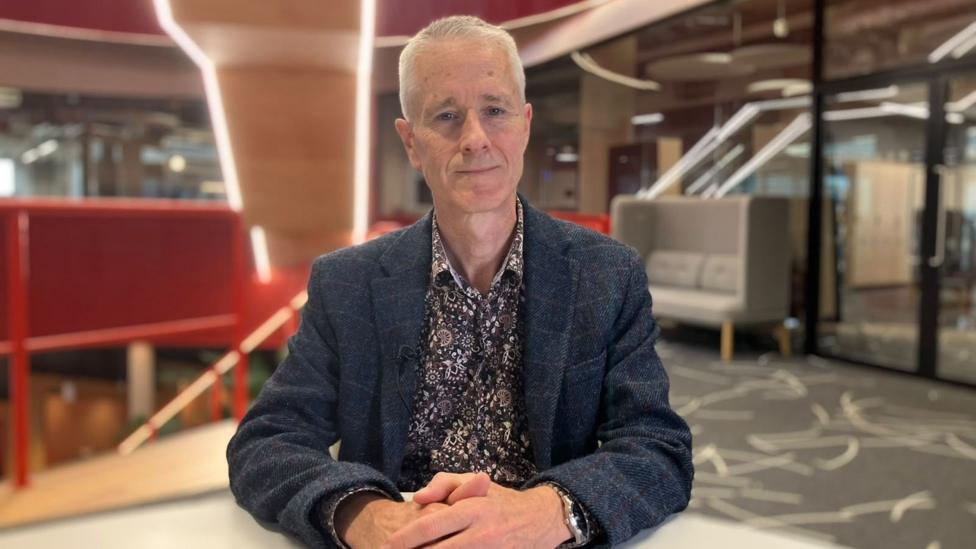 Jonathan Scourfield is wearing a patterned shirt and dark blazer. He is sitting in a modern office building. Jonathan is looking straight at the camera. He has short spiky hair and blue eyes.