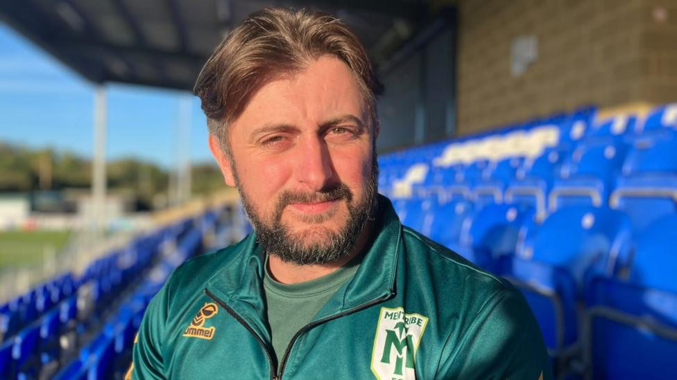 Callum Tipton sits in a football stadium stand wearing a dark green quarter-zip sports top with the “MENTRIBE FC” crest on the chest. Callum has medium length brown hair with short sides and has a short beard.
