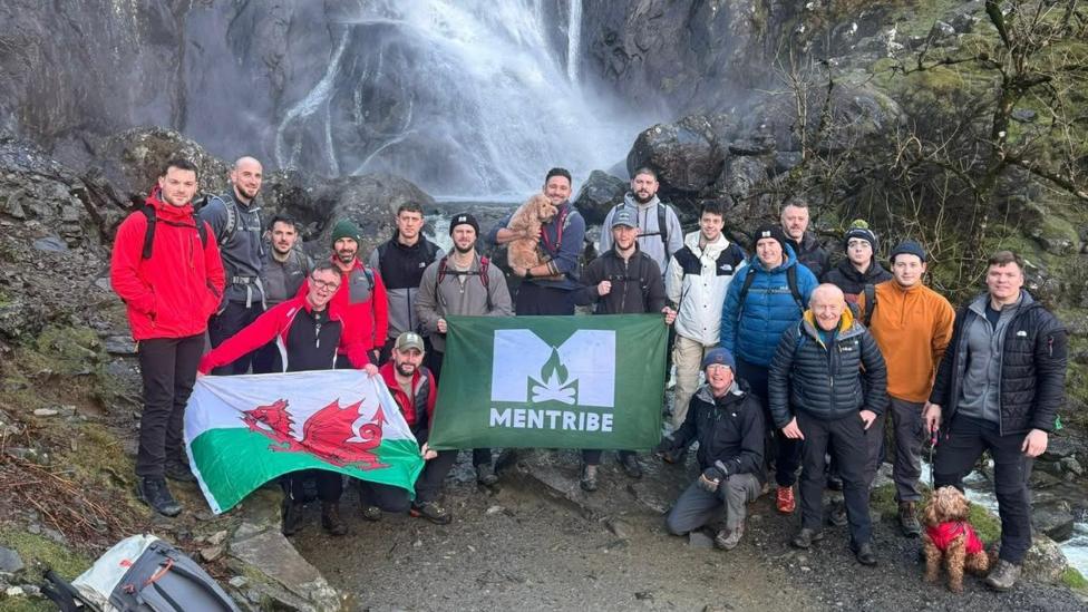 A large group of hikers pose at the base of a waterfall cascading down a steep rocky cliff. The group stands on a rocky path surrounded by mossy rocks and bare trees, with several people holding a Welsh flag and a green “MENTRIBE” banner. Two dogs are visible, one being held by a participant and another wearing a red coat near the front. Backpacks rest on rocks in the foreground, while mist from the waterfall rises behind the group in an outdoor mountain setting.