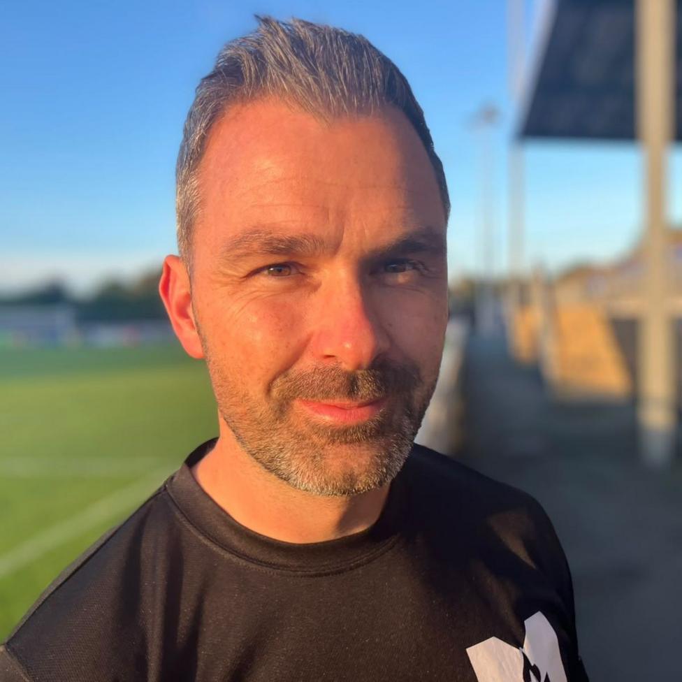 Close-up portrait of Gavin Roberts standing beside a football pitch in warm evening sunlight. Gavin is wearing a black sports top, with the background softly blurred to show part of a football field on the left. Gavin has short grey hair with a spikey fringe. He also has a short, grey beard.