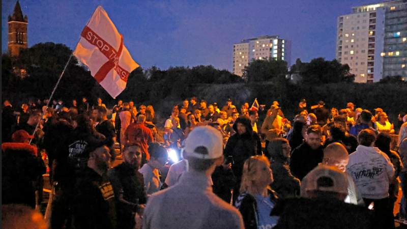 Demonstrators are standing around, it's quite dark and there are a couple of tall buildings in the background. A red and white flag reads Stop the Boats