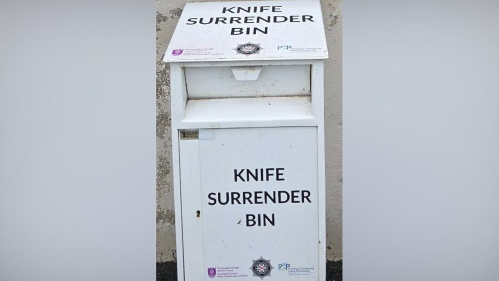 A white bin has "KNIFE SURRENDER BIN" in the centre in black font