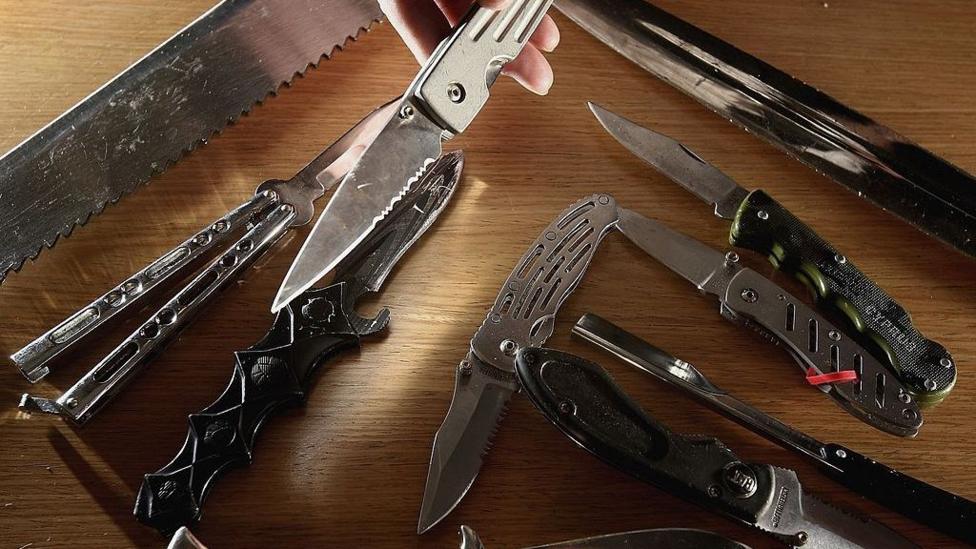 Approximately 10 black and silver knives, of different variations sit on a wooden table.
