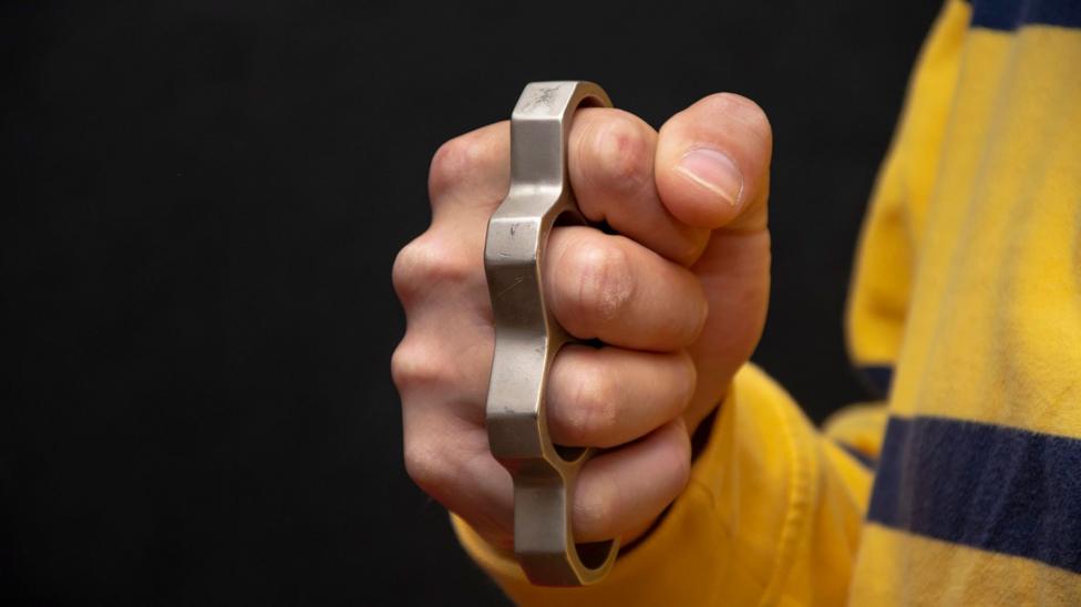 A man wearing a yellow and navy rugby top has a silver knuckle-duster on his right hand.