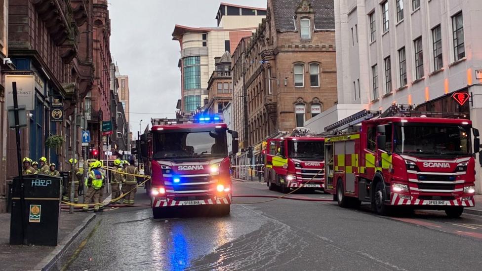 Fire engines parked in a city centre street with firefighters standing beside one of them