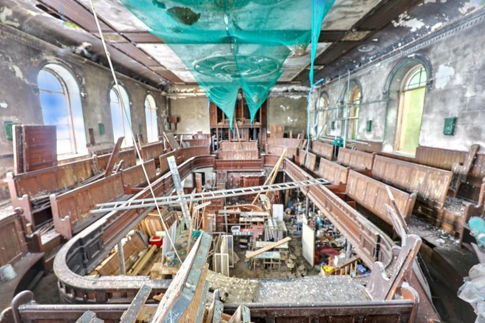 An inside view of the church with curved rows of wooden pews, tall arched windows and grey mottled peeling walls. Debris, ladders and building materials cover the lower floor and torn green netting hangs from the crumbling ceiling.