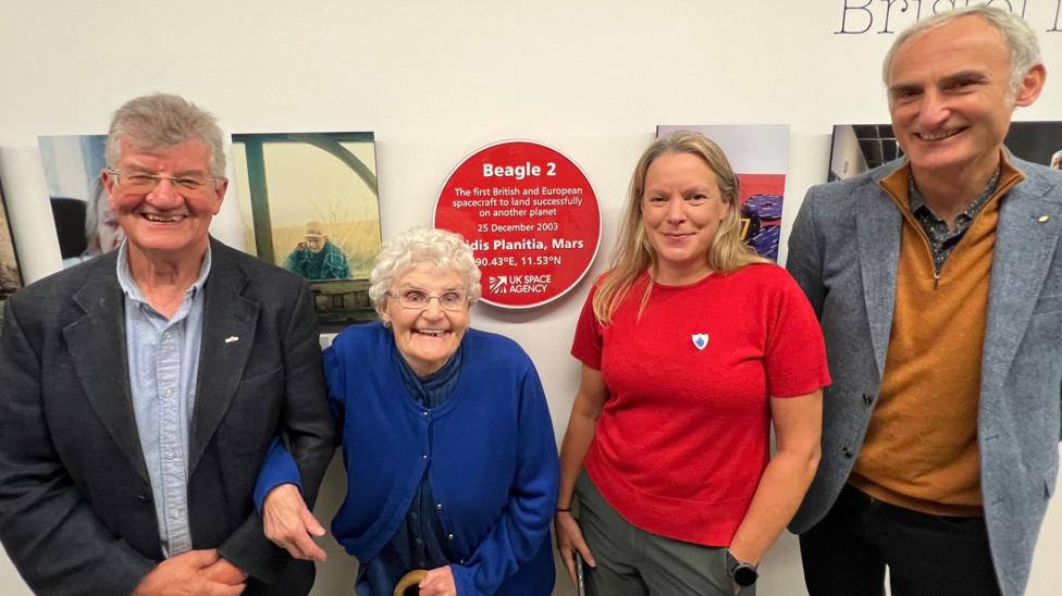 A red plaque that says Beagle 2 with the coordinates of the landing on Mars hangs on a white wall next to a series of pictures. Four people stand posing for a photo next to the plaque, including a happy-looking older woman and a smiling younger woman in a red T-shirt.