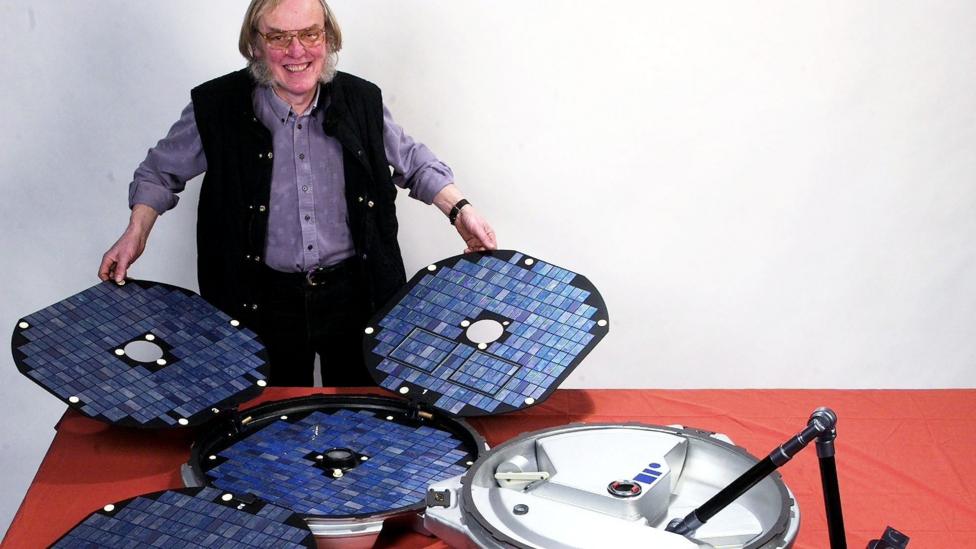 A man wearing a purple shirt and black sleeveless coat holds open the Beagle 2 probe, comsisting of four solar panels that open up and a round dish.