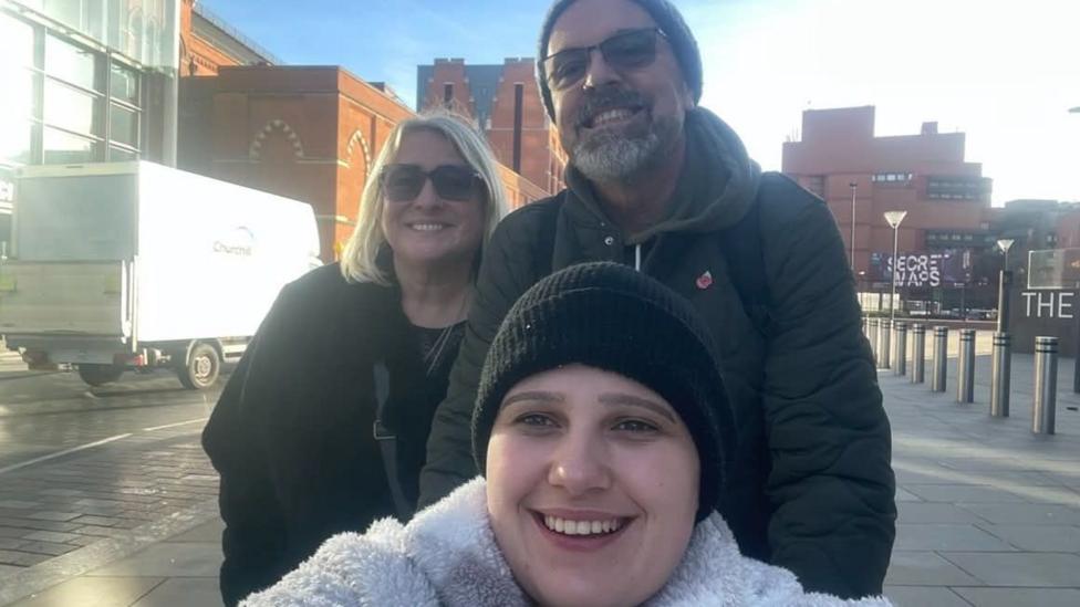 A selfie of Phoebe with both her parents during a trip to London. Phoebe is wearing a black beanie hat and white fluffy jumper. She is taking the selfie, with both her parents stood behind her. Her dad is wearing a dark green coat with a hood, a grey beanie hat and sunglasses. Her mum has blonde shoulder length hair and is wearing a black coat. They are all smiling and looking at the camera.