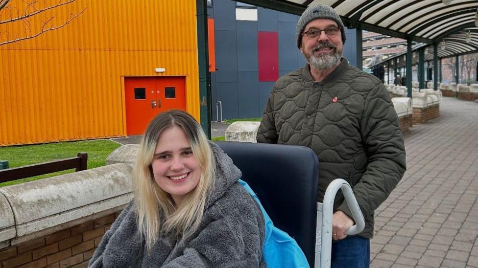 Phoebe is wearing a grey hoodie. She is smiling and looking at the camera. She is outside and is being pushed in her wheelchair by a man wearing a beanie and glasses behind her.
