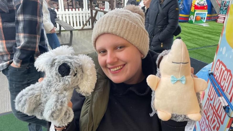 Phoebe is photographed whilst on a family trip to London. She is holding two plush toys she had won as a prize. One is a grey koala bear (left) and the other is a hedgehog with grey fur and a yellow stomach and face. A blue bow tie is attached under its face. Phoebe is holding the plush toys up to her face. She is smiling and looking at the camera. She is wearing a beige beanie hat, black hoodie and dark green body warmer.