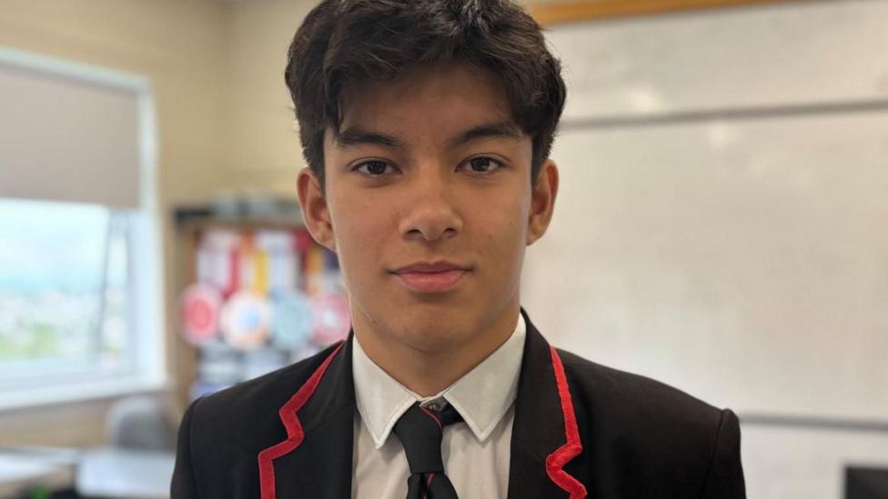 Pupil Tomo in school uniform standing in a classroom, with a whiteboard, desks and digital display screen in the background.