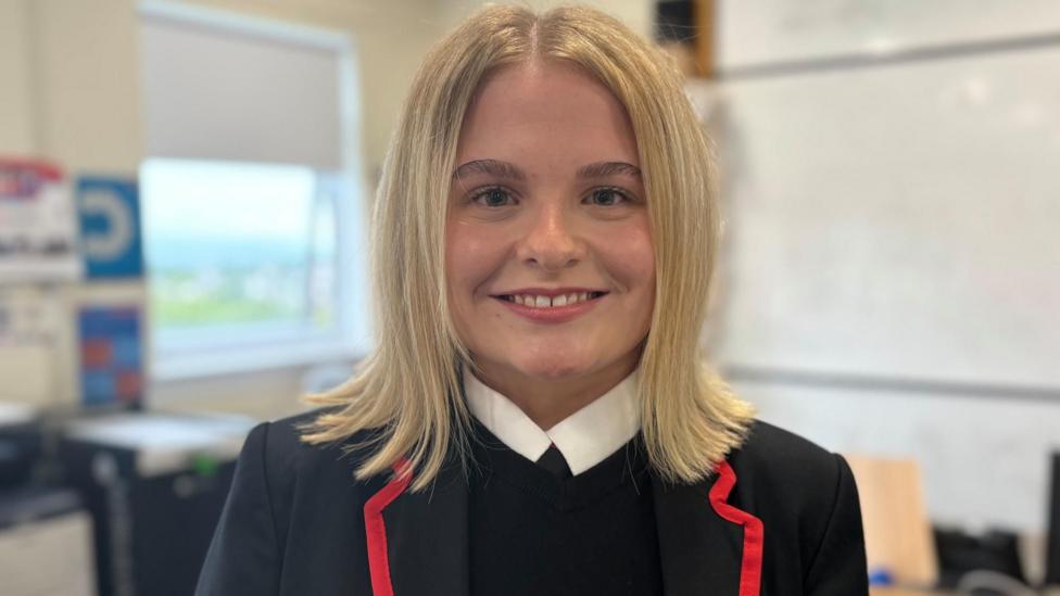Pupil Anna in school uniform standing in a classroom, with a whiteboard and teaching equipment visible in the background.