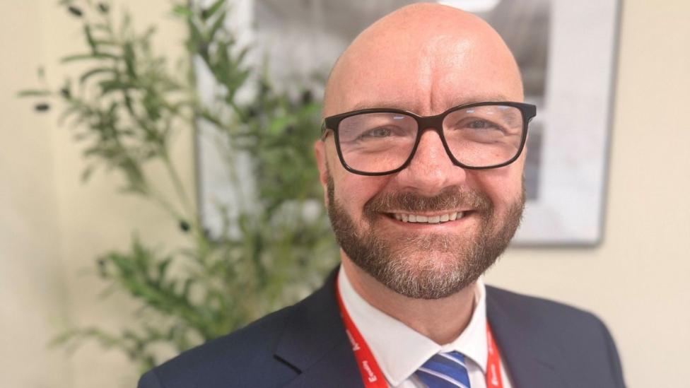Stuart Clark is wearing glasses, a dark suit jacket, white shirt and blue striped tie, with a red lanyard around his neck. The photograph is taken indoors with blurred green plants and framed wall art in the background.